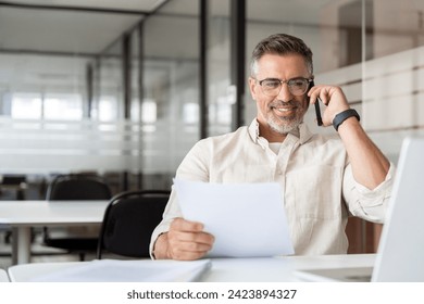 Middle aged Latin businessman having call on smartphone with business partners or clients. Smiling mature Hispanic man sitting at table talking by mobile cellphone, using laptop, paper documents - Powered by Shutterstock