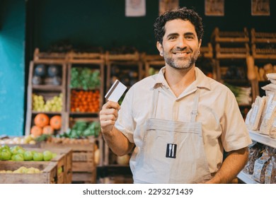 Middle aged latin greengrocer looking at camera while holding credit card in hand. Copy space. - Powered by Shutterstock