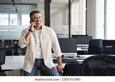 Middle aged Latin or Indian businessman having call on smartphone with business partners or clients. Smiling mature Hispanic man in relaxing pose talking by mobile cellphone at work in modern office.  - Powered by Shutterstock