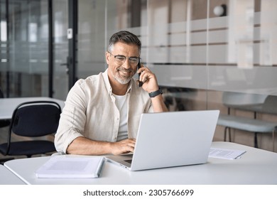 Middle aged Latin or Indian businessman having call on smartphone with business partners or clients. Smiling mature Hispanic man sitting at table talking by mobile cellphone at workplace in office.  - Shutterstock ID 2305766699