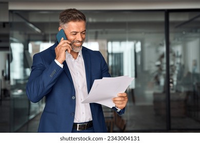 Middle aged Latin or Indian businessman walking, having call on smartphone with business partner and looking at documents. Mature Hispanic man talking by mobile cellphone at work in office, copy space - Powered by Shutterstock