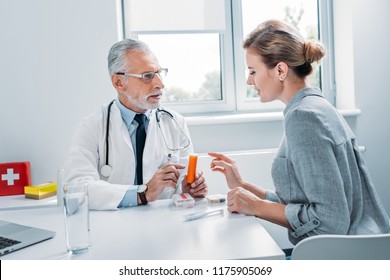 Middle Aged Male Doctor Pointing At Pills To Female Patient At Table In Office 