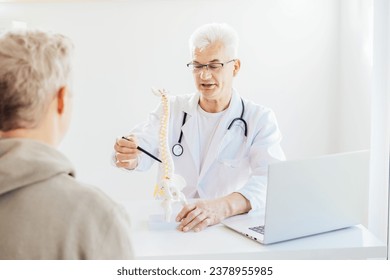 Middle aged male doctor pointing on human spine model with a pen and explaining patient his problem in medical clinic. Spinal healthcare and back pain treatment concept. Selective focus, copy space - Powered by Shutterstock