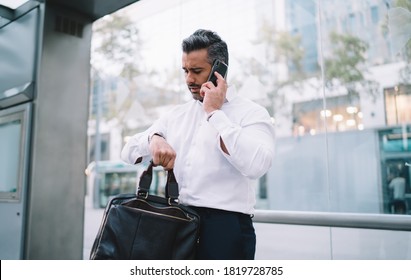 Middle aged male employee checking wrist watch time while waiting bus on stop calling to colleague for to warn about delay to business meeting,mature boss communicating via mobile during daily routine