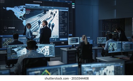 Middle Aged Male and Female Government Officers Discuss Working Matters next to a Big Digital Screen with Satellite Surveillance Footage with Face Recognition Software. Police Investigators at Work.