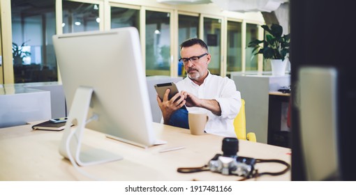 Middle aged male manager watching uploaded photos on tablet sitting at desk with computer photo camera and mug with coffee on break