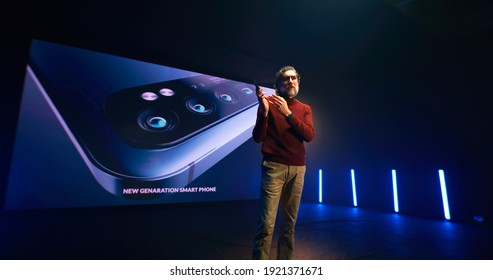 Middle Aged Male Speaker Demonstrating Brand New Mobile Phone And Talking During Presentation On Stage Against Big LED Screen