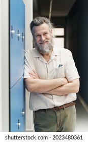 Middle Aged Male Teacher With Beard Standing In School Hallway With Blue Lockers. Teaching And Education Vocational Profession