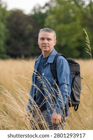 Middle aged man with backpack standing in tall dry grass. Outdoor adventure, hiking, freedom, travel and peaceful nature lifestyle concept.