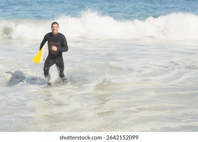 Middle aged man in black wetsuit emerging from ocean at beach carrying white and yellow surfboard. Adventure, sport, aquatic, active, exploration, outdoors, energetic