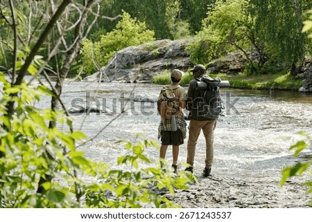 Middle aged man and boy standing on rocky riverbank fishing together, both wearing backpacks, surrounded by lush green trees and flowing water in outdoor nature setting
