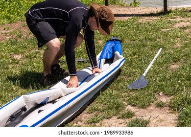 Middle Aged Man In Brown Hat, Black Swim Shirt And Shorts, Folding Up Deflated Inflatable Kayak. Preparing It For Storage.  Paddle On The Ground Next To Kayak. 