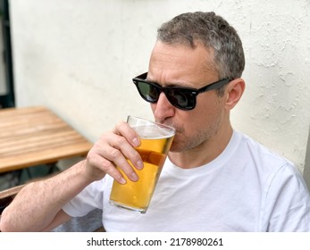 Middle Aged Man Is Drinking Beer In A Cafe Or Restaurant Pub Outdoor. Drinking Alcohol. Candid. Refreshment, Summer Time, Pub Garden. Wearing White T-shirt And Sunglasses. Drinking Lager Or Ale Beer