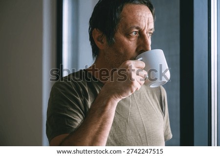 Middle aged man drinking coffee from a cup while standing by a window, thoughtful mood, natural daylight, quiet indoor lifestyle moment. Selective focus.