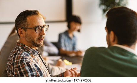 Middle Aged Man In Eyeglasses And With Beard Talking To Colleague Sitting Next To Him At Desk During Business Meeting