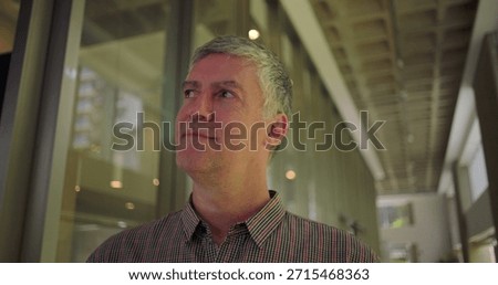 Middle aged man glancing to the side with subtle smile in modern office hallway thoughtful relaxed and optimistic expression in professional environment

