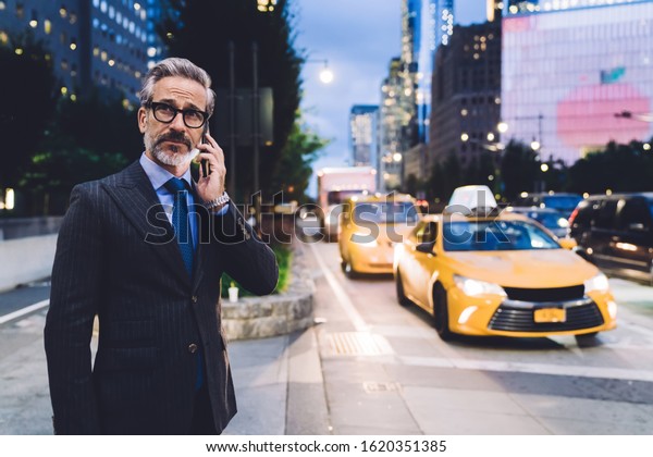 Middle aged man in glasses and business suit talking
on smartphone and standing against evening New York road while
looking away
