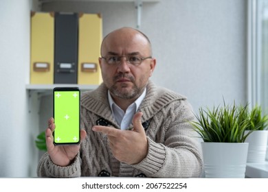 Middle Aged Man With Glasses Shows Off Something On Smartphone With Green Screen To Camera. Chroma Key Phone Set Up For Advertising. Serious Businessman Holding Mobile Phone. Selective Focus.