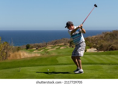 Middle Aged Man Golfing, Caught Mid-swing Off The Tee Box. On Ocean Side Tropical Golf Course With Deep Blue Sea And Clear Blue Sky. Copy Space.