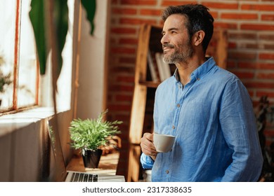 A middle aged man is having a coffee break at home while he's working and looking through the living room window with a smile on his face - Powered by Shutterstock