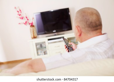 Middle Aged Man In Home Bright Living Room Sitting On Comfortable Couch And Watching TV