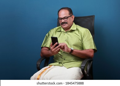 Middle Aged Man Of Indian Origin Sitting On A Chair  Checking His Mobile Phone