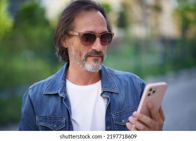 Middle Aged Man Looking At Smartphone Standing On Street While Enjoying Free Time Off Work. Mature Man Read Travel Digest Using Mobile Phone Traveling Old Town. Business On The Go. 