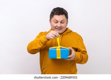 Middle Aged Man Opening Gift Box, Unwrapping Birthday Present For Celebrating Holiday, Pulling Ribbon, Looking At Gift, Wearing Urban Style Hoodie. Indoor Studio Shot Isolated On White Background.