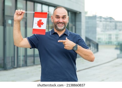 Middle Aged Man Points To The Canadian Flag In The Background Of The City. New Life In Canada.