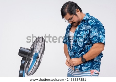 Middle aged man seeking relief from the heat by unbuttoning his Hawaiian shirt in front of an electric fan, isolated on white. [[stock_photo]] © 