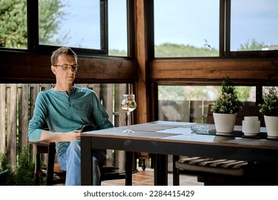 Middle aged man sits on the veranda of country house with a glass of wine and uses smartphone - Powered by Shutterstock