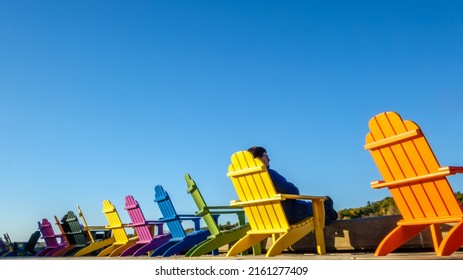 Middle Aged Man Sitting In A Row Of Brightly Colored Adirondack Chairs Along Waterfront In Fall With Fall Foliage In The Distance.