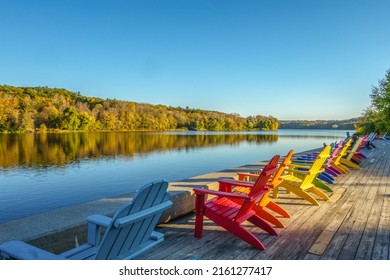 Middle Aged Man Sitting In A Row Of Brightly Colored Adirondack Chairs Along Waterfront In Fall With Fall Foliage In The Distance.