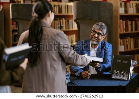 Middle aged man sitting at table handing book to young adult Asian woman during book presentation event in library, bookshelves filled with books in background