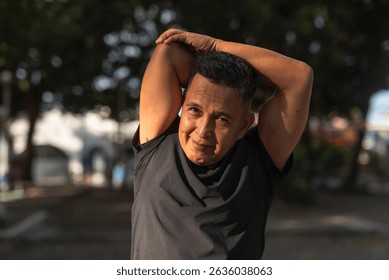 A middle aged man smiling while doing a tricep stretch, preparing for exercising at the park. Fitness in 50s. - Powered by Shutterstock - Get 15% off with code: PIKWIZARD15