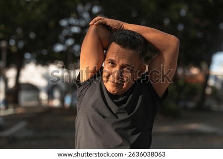 A middle aged man smiling while doing a tricep stretch, preparing for exercising at the park. Fitness in 50s.