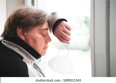 A Middle Aged Man Stands At The Window Looking At An FFP 2 Mask. He Is Worried About The Uncertain Future Of Society Due To The Corona Pandemic. Not A Typical Model But A Normal 50 Year Old Man.