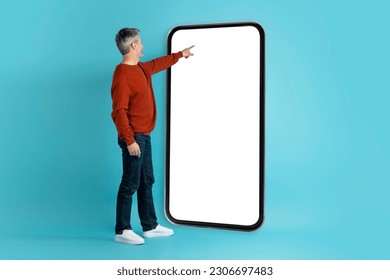 Middle aged man travel enthusiast touching white empty screen of huge cell phone displaying travel booking app. Tourist buying tickets, making online hotel reservation, blue studio background, mockup