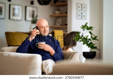 Middle aged man using cellphone and relaxing in an armchair at home. Confident male wearing turtleneck sweater and drinking tea. 