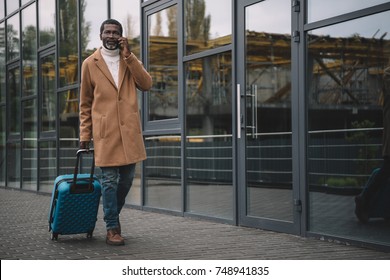 Middle Aged Man Walking On A Street With A Wheeled Bag And Talking By Smartphone