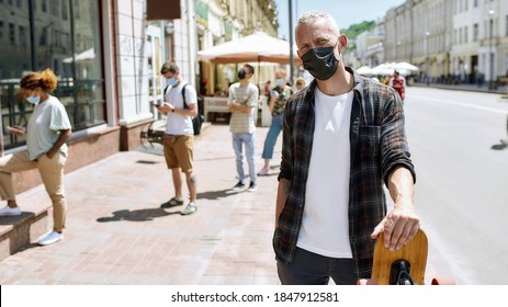 Middle aged man wearing mask holding a longboard and looking at camera. People waiting in line to collect their orders, purchases from the pickup point during coronavirus lockdown in the background