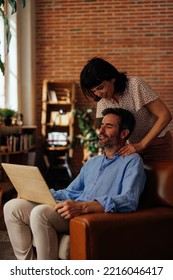 A Middle Aged Man Is Working Online At Home Sitting In An Armchair While His Wife Behind Him Giving Him A Back Rub.