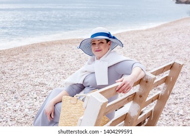 Middle Aged Mature Woman Posing On The Beach In The Sea. Wearing Grey Dress, Straw Hat And Bag