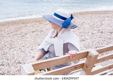 Middle Aged Mature Woman Posing On The Beach In The Sea. Wearing Grey Dress, Straw Hat And Bag