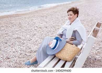 Middle Aged Mature Woman Posing On The Beach In The Sea. Wearing Grey Dress, Straw Hat And Bag