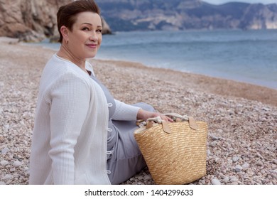 Middle Aged Mature Woman Posing On The Beach In The Sea. Wearing Grey Dress, Straw Hat And Bag