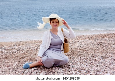 Middle Aged Mature Woman Posing On The Beach In The Sea. Wearing Grey Dress, Straw Hat And Bag