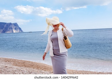 Middle Aged Mature Woman Posing On The Beach In The Sea. Wearing Grey Dress, Straw Hat And Bag