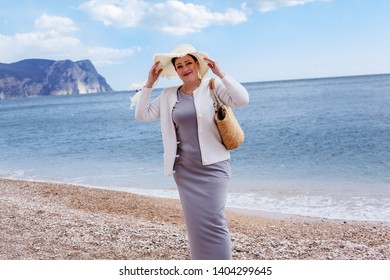 Middle Aged Mature Woman Posing On The Beach In The Sea. Wearing Grey Dress, Straw Hat And Bag