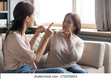 Middle Aged Mother And Adult Daughter Speaking Sign Language, Sitting On Couch In Living Room At Home, Young Woman And Serious Mature Mum Chatting, Communicating, Two Generations